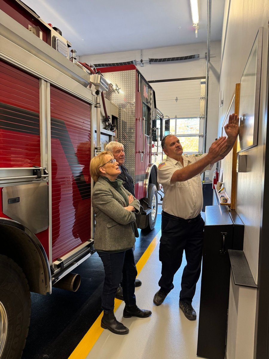 Inside a fire station, a uniformed firefighter gestures toward a wall display while a woman and an older man watch. A large red fire engine is parked beside them. There is text visible on the fire engine and wall display.