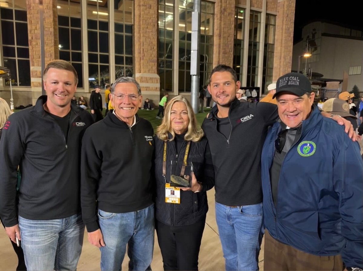 Five adults pose together for a nighttime group photo outside a large building. The woman in the middle holds a small award or keepsake, and several people wear jackets with visible logos or lettering.