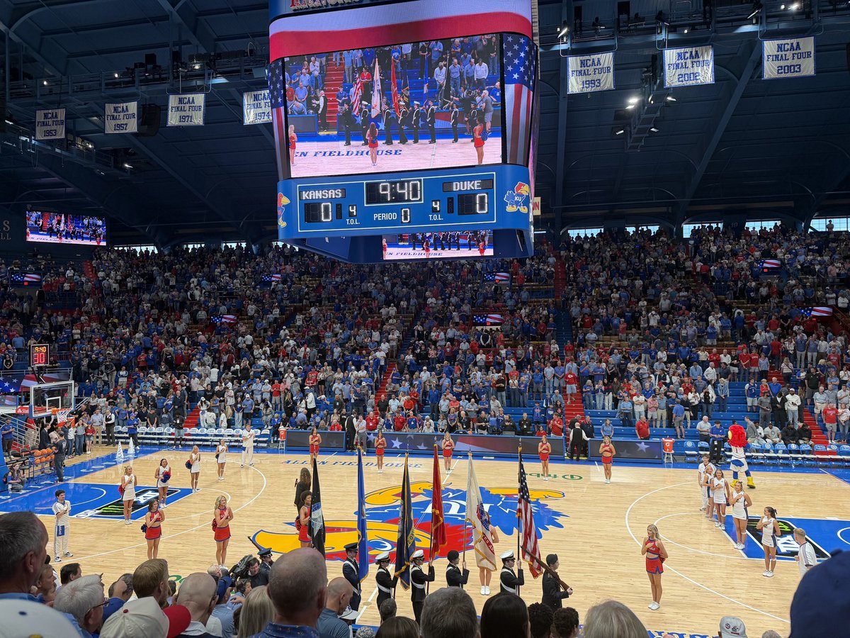 A packed indoor basketball arena during a game or pregame ceremony. Cheerleaders stand around the court, and uniformed flag bearers hold several flags near center court. A large overhead video scoreboard shows Kansas and Duke with a game clock. Banners hang from the rafters, and text appears on the scoreboard and banners.