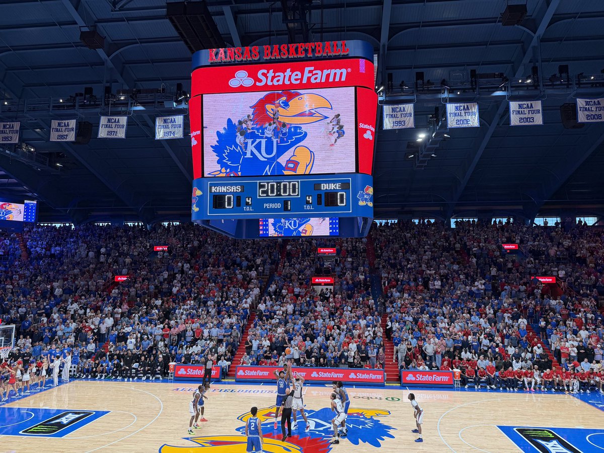 A college basketball game is underway in a packed indoor arena. Players gather at center court for the opening tip beneath a large illuminated scoreboard. Banners hang from the rafters, and there is text throughout the scene.