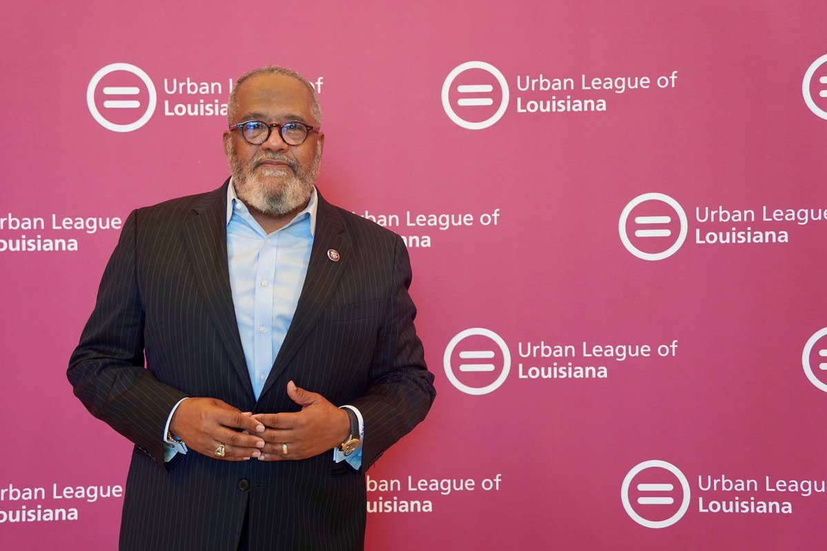 A man wearing glasses, a dark pinstriped suit, and a light blue shirt poses in front of a pink step-and-repeat backdrop. The backdrop contains repeated logos and text.