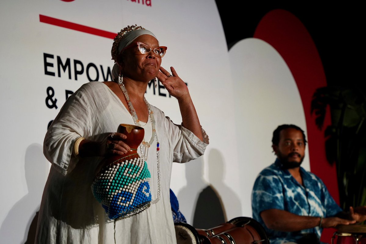 A woman in white clothing, glasses, and a headwrap stands on a stage holding a wooden cup and raising one hand. A beaded bag hangs at her side, and a seated man with drums is beside her. A backdrop with large text and red-and-white graphics is behind them.