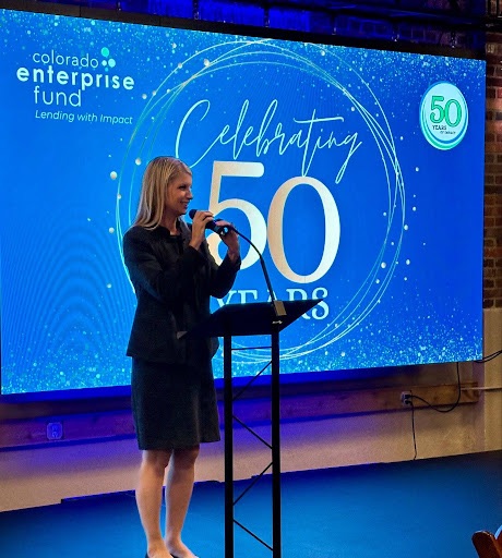 A woman speaks at a lectern in front of a large blue anniversary display. The scene appears to be an indoor event, with stage lighting and brick walls. There is text on the display.