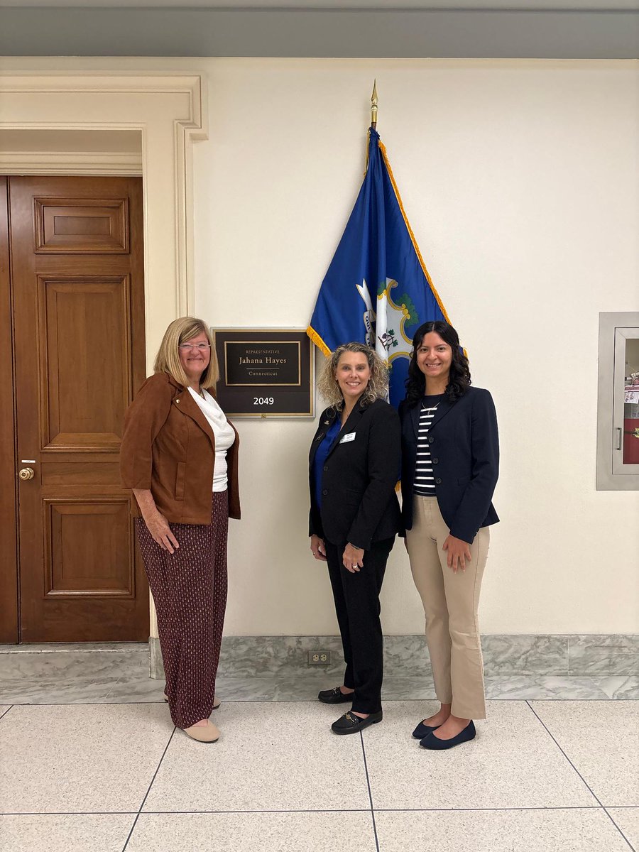 Three women stand together in a formal hallway beside an office door, a wall plaque, and a blue state flag. Text is visible on the plaque.