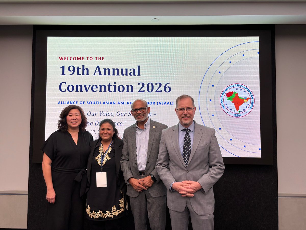 Four adults pose for a photograph in front of a large screen displaying a convention welcome slide with a logo and other text.
