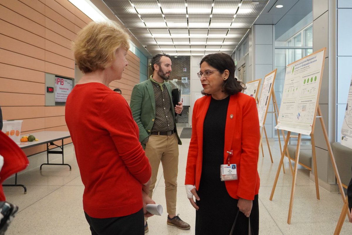 Three people talk in a bright indoor hallway beside poster displays and a table with small items. The posters and signs contain text.
