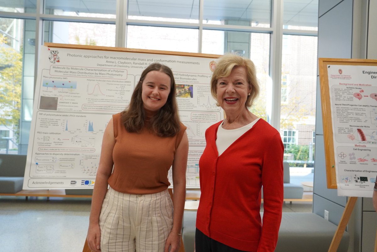 Two smiling women pose indoors in front of academic research posters. The posters contain text, charts, and diagrams.