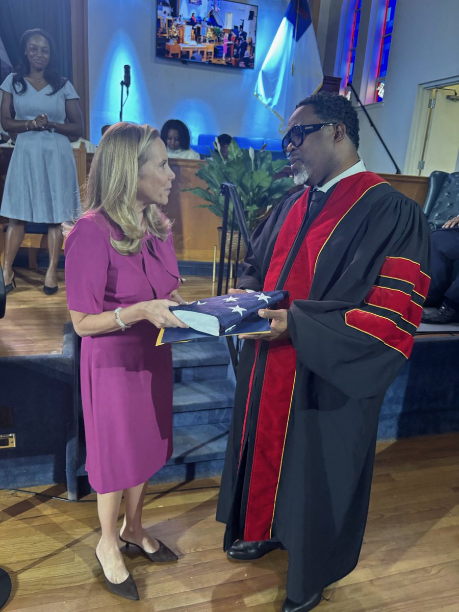 A woman in a magenta dress hands a folded American flag to a man wearing academic regalia inside a church or auditorium. Other people, stage seating, a plant, and a wall-mounted screen are visible in the background. There is text on the screen, but it is not legible.