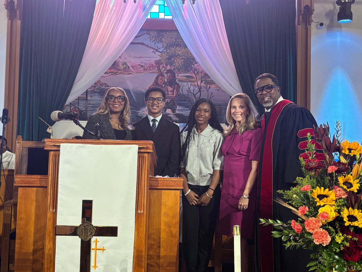 A group of five people pose together at the front of a church, beside a wooden pulpit. One person wears clerical robes, and a large arrangement of flowers stands nearby. Drapes and a religious mural decorate the background. No readable text is visible.
