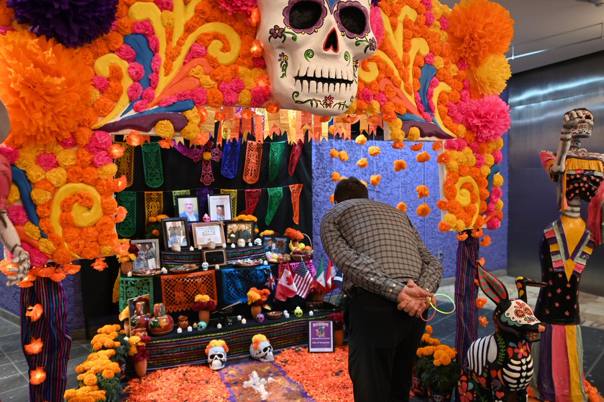 A colorful Day of the Dead altar decorated with flowers, papel picado, skulls, candles, offerings, and framed photographs. A person stands in front of it, with decorative skeleton figures nearby. There is some text on a small sign.