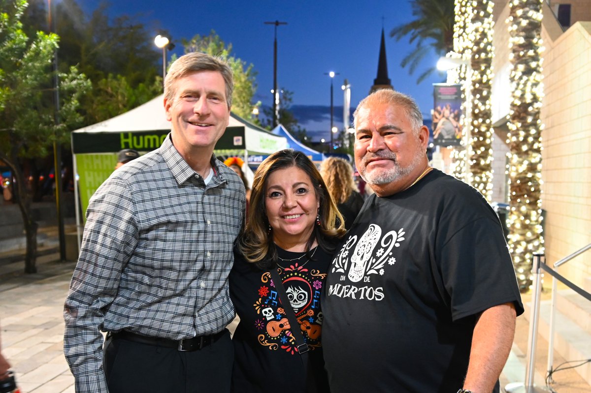 Three adults pose together for a photo at an outdoor evening event. Behind them are event tents, people, trees, and palm trees wrapped in lights. There is text on the tents and on two people’s shirts.
