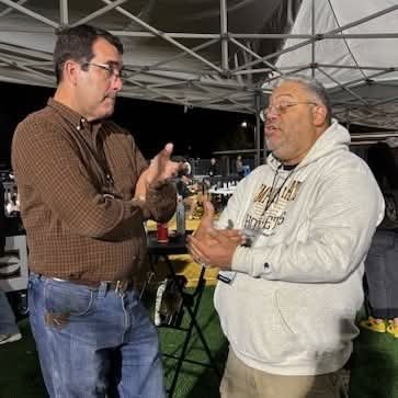 Two men are talking beneath a canopy at an outdoor nighttime gathering. Tables, chairs, and other people are visible in the background. There is text on one man's hoodie.
