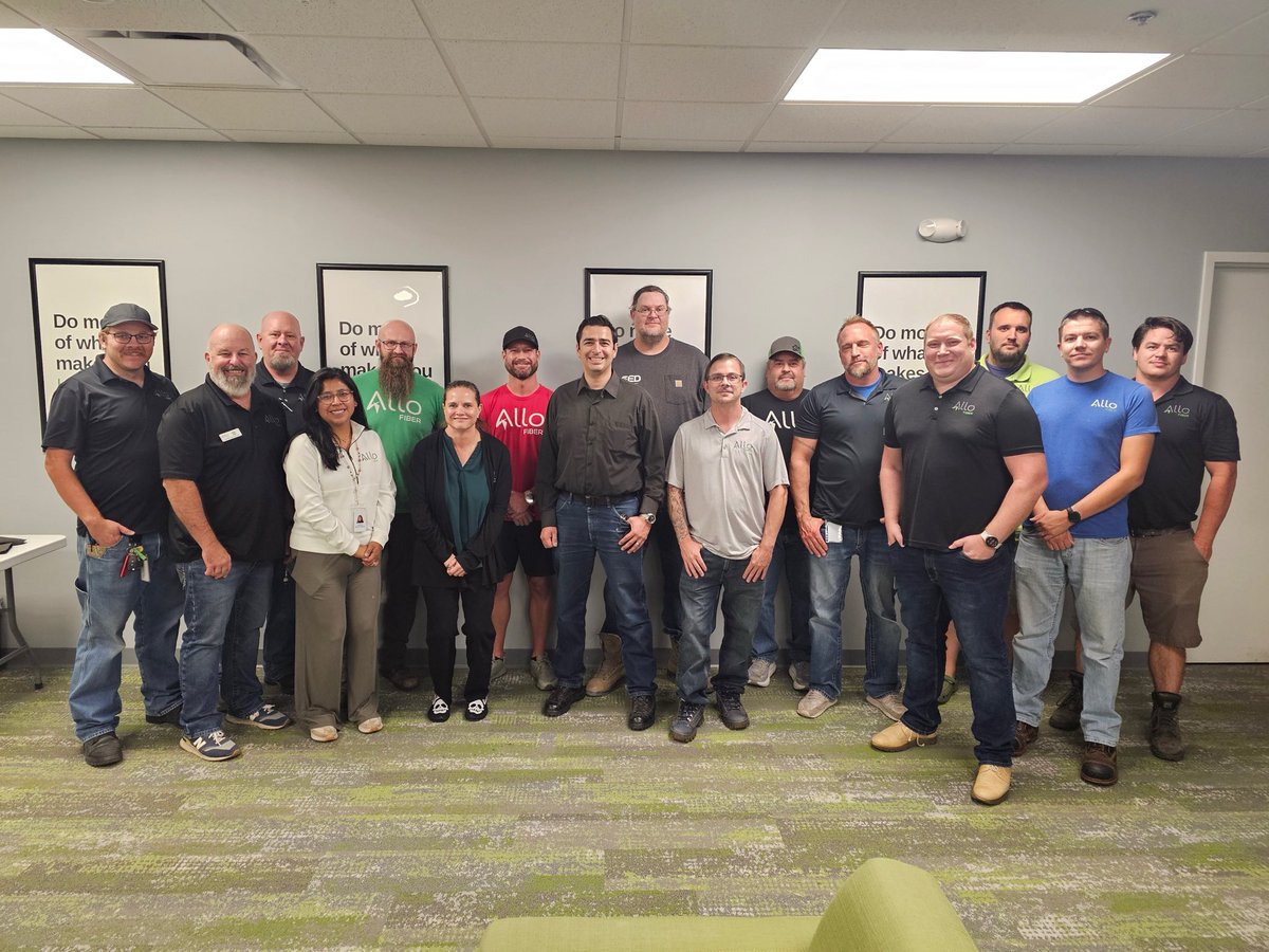 A group of people pose together in an office or meeting room. Several wear branded company shirts, and framed posters are visible on the wall behind them.
