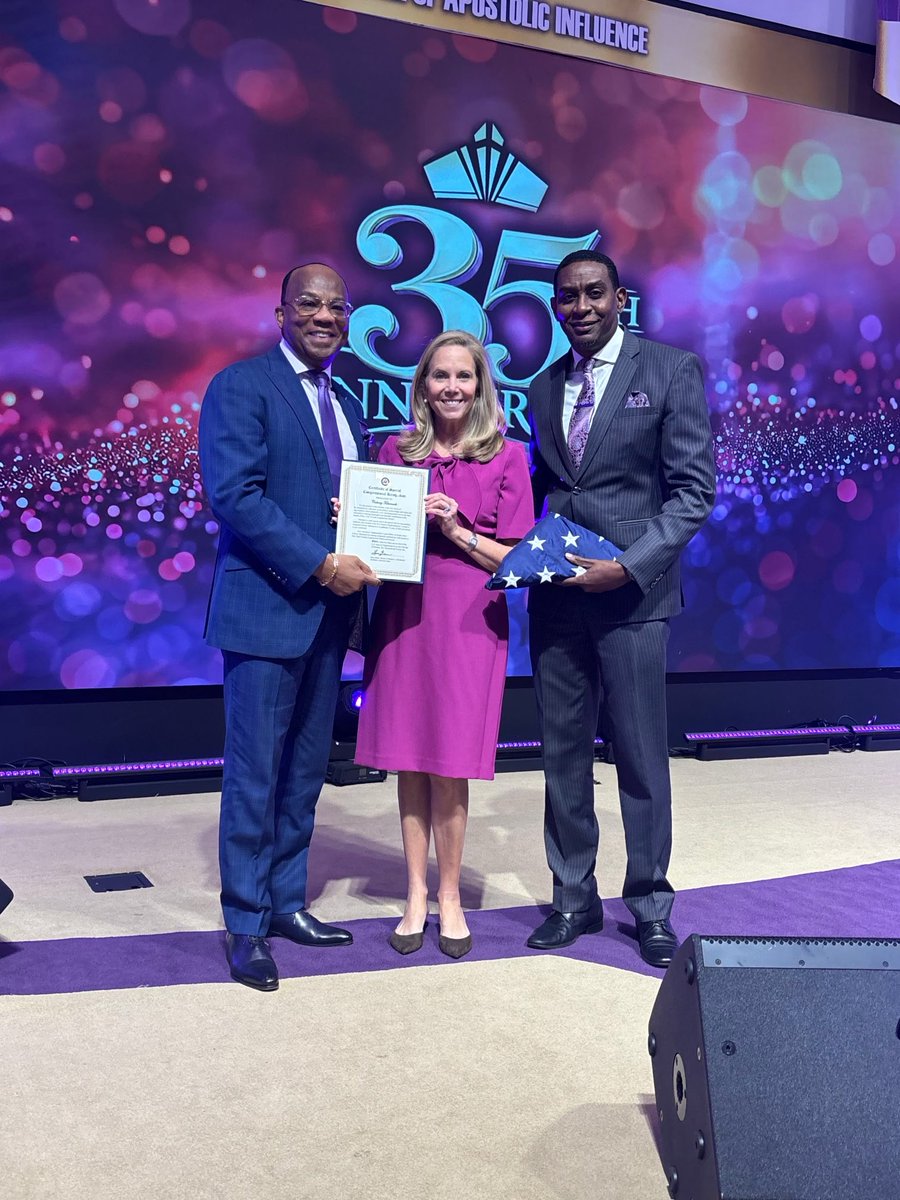 Three formally dressed people stand together on a stage at an event. The woman in the middle holds a framed certificate, while the man on the right holds a folded American flag. A large colorful event backdrop with prominent lettering is behind them.