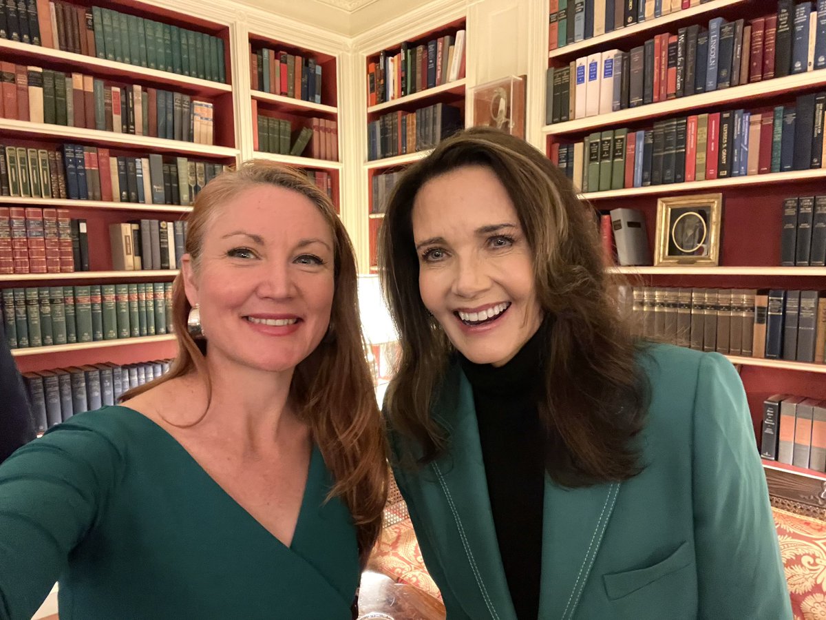A selfie of two smiling women standing together in a warmly lit room lined with bookshelves. There is text on the book spines.