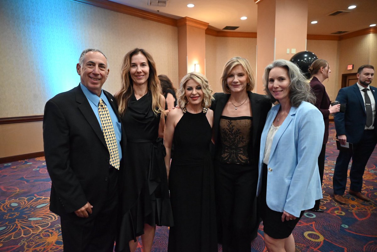 A group of five adults pose together for a photograph at an indoor formal event. Other attendees and a black balloon are visible in the background. There is a small amount of visible text in the room.