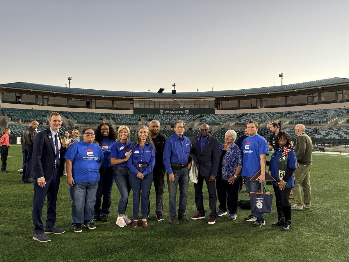 A group of people pose for a photo on the field of a soccer stadium, with rows of empty seats and the stadium structure behind them. Some people wear matching blue shirts, and one person holds a bag. Text is visible on clothing and in the stadium.