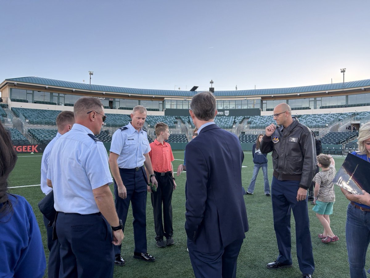A group of adults and children stand and talk on the grass inside a large, mostly empty football stadium. Several men wear light-blue uniforms, while others wear suits or casual clothing. Text is visible on stadium signage and clothing.