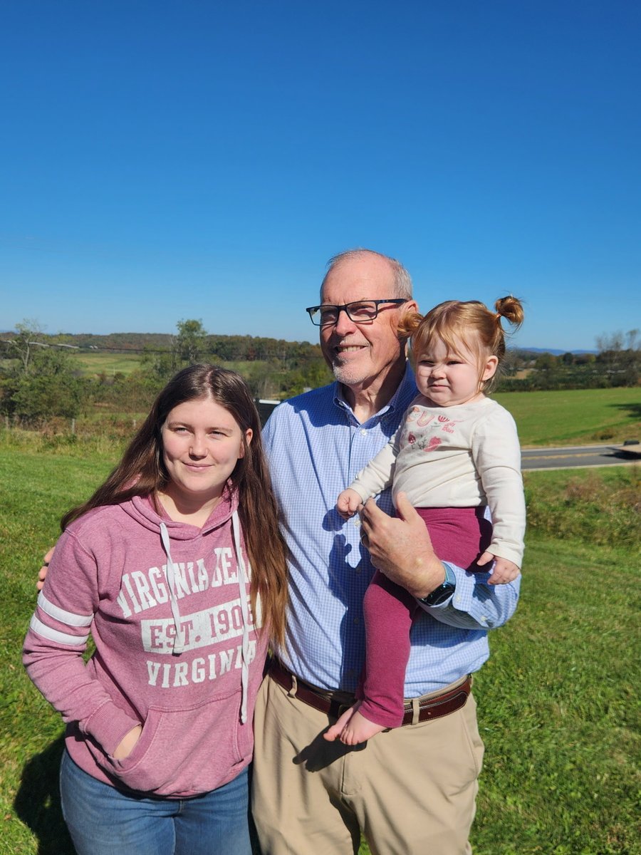 A family photo outdoors on a sunny day, showing an older man holding a toddler beside a teenage girl. They are standing in a grassy field with a road and trees in the background. Text appears on the girl's sweatshirt.