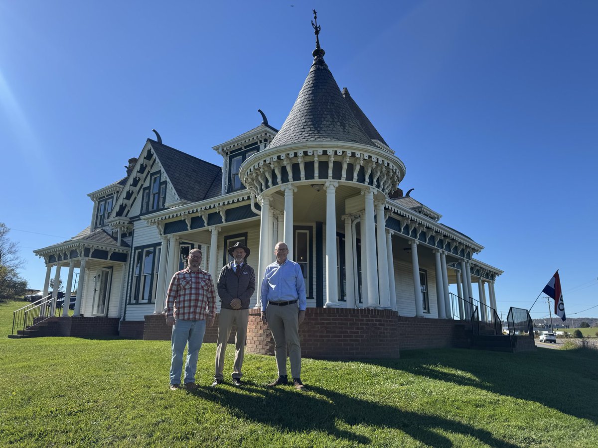 Three men pose on a lawn in front of a large ornate Victorian-style house with a turret and wraparound porch, beneath a clear blue sky. A flag is visible near the roadside. There is text on the flag.
