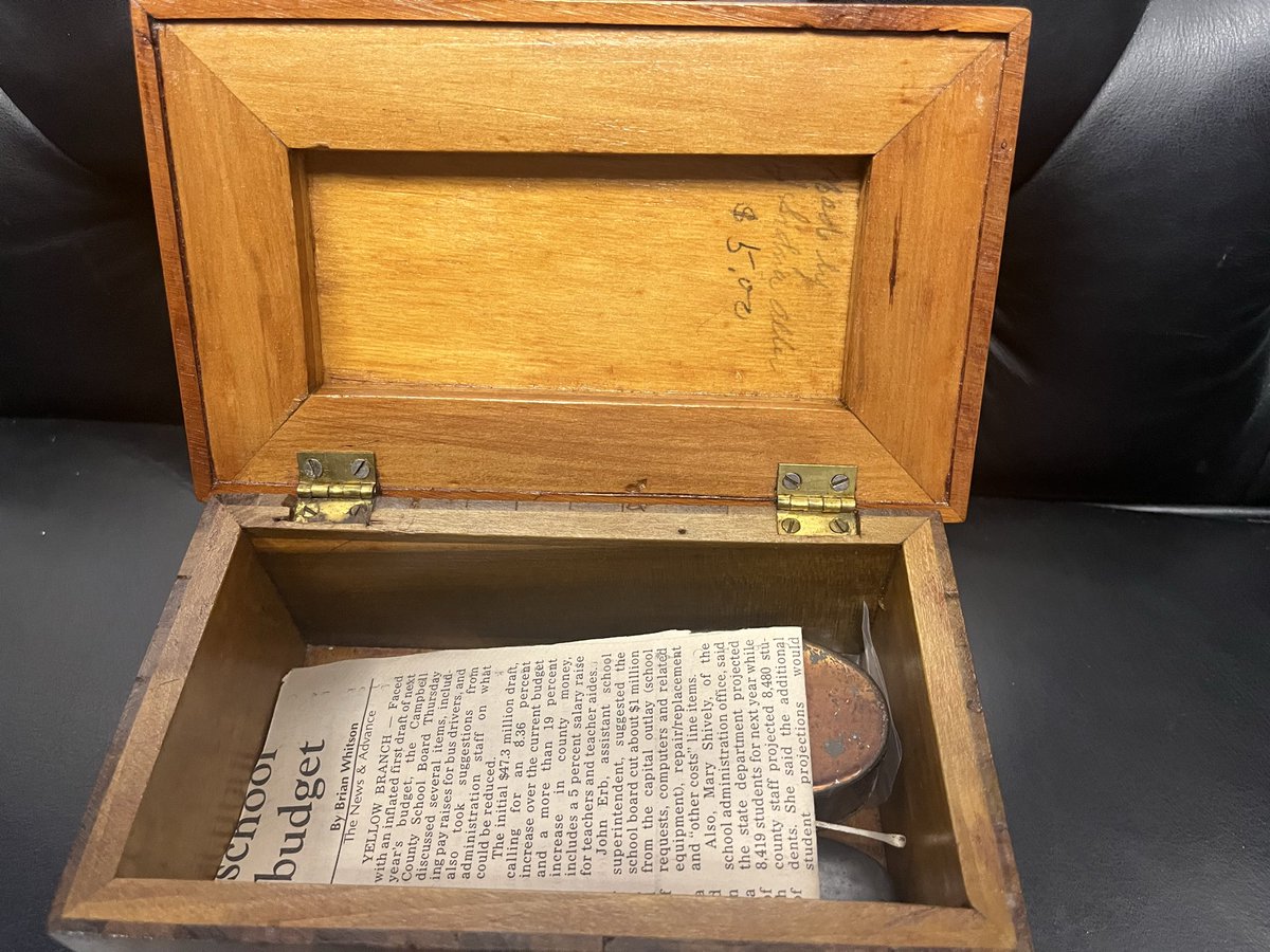 An open wooden box with brass hinges sits on a dark surface. A folded newspaper and a small, rusty metal object are inside. Faint handwriting is visible on the inside of the lid, and printed text appears on the newspaper.