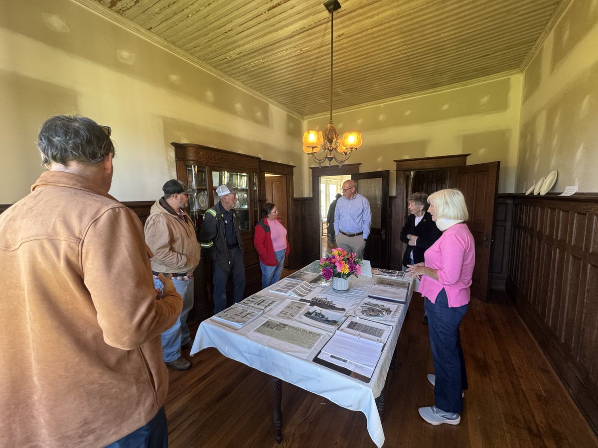 A group of people stand around a table displaying documents and photographs in a wood-paneled room. A vase of colorful flowers sits among the materials, and a chandelier hangs from the ceiling. Text is visible on some of the documents.