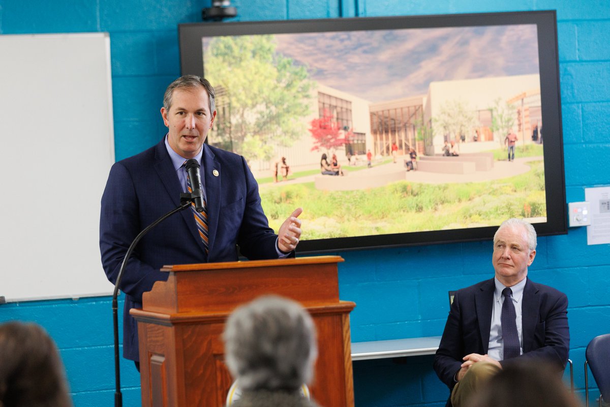 A man in a suit speaks at a wooden podium with a microphone while gesturing to an audience. Another suited man is seated nearby. A large display behind them shows an architectural rendering of a modern building and grounds. The scene appears to be indoors in a room with bright blue walls. Some text is visible on a notice at the right.