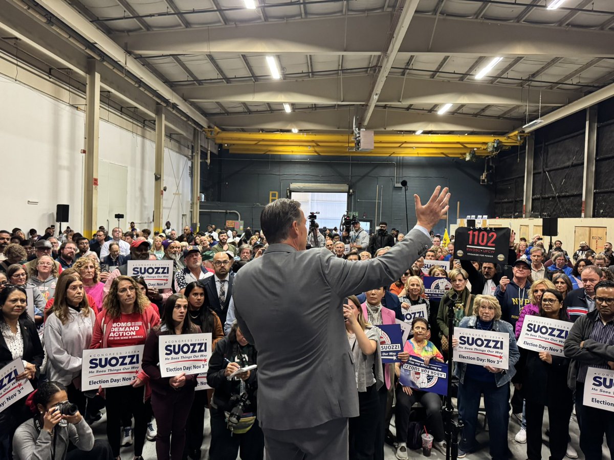 A man in a gray suit addresses a large crowd inside an industrial hall, raising one hand as people hold campaign signs. Text appears on the signs and on a shirt.