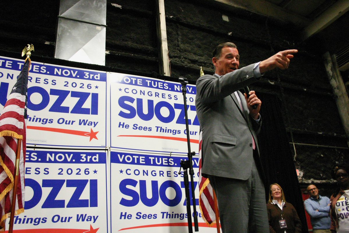 A man in a gray suit speaks into a microphone and points toward the audience at a political campaign event. Campaign signs and American flags are visible behind him, with several people watching. There is text on the signs.
