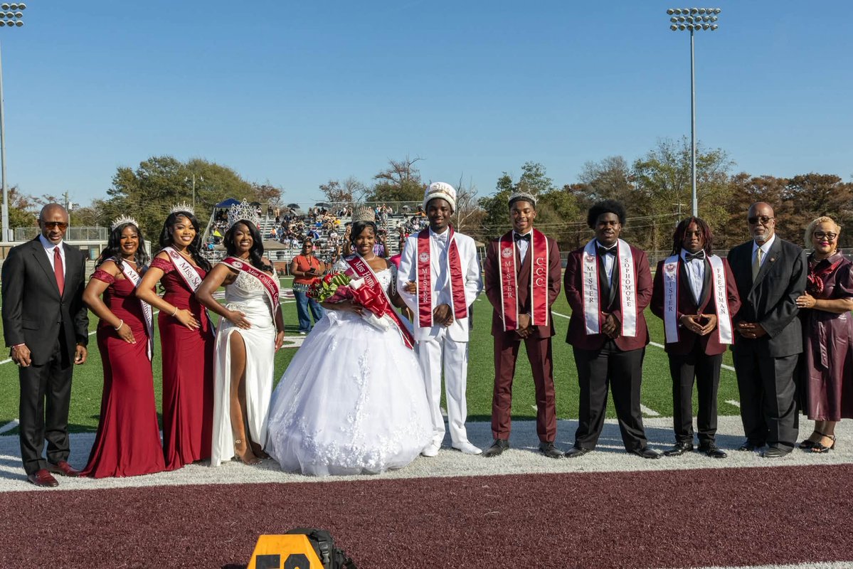 A group of students and adults pose together on a football field or track. Several students wear formal outfits and ceremonial sashes, and one wears a white gown and holds flowers. Bleachers, spectators, trees, and stadium lights are visible in the background. There is text on the sashes.