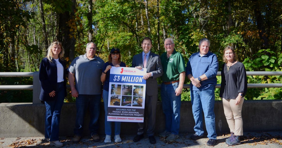 A group of eight adults pose outdoors in front of a wooded area, holding a poster announcing funding for a regional flood mitigation project. Text appears on the poster.