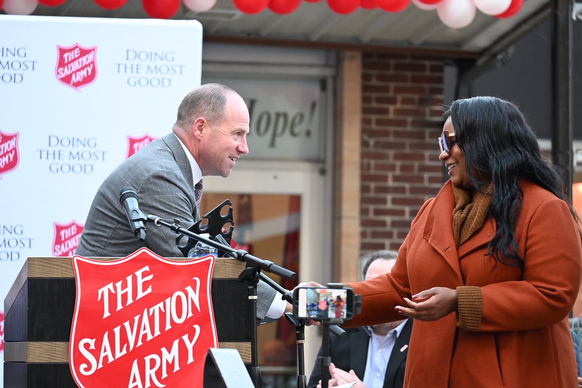 A man speaking at a podium and a woman in an orange coat interacting at an outdoor Salvation Army event. Microphones, a phone on a stand, red and white balloons, and event signage are visible. The image contains text.