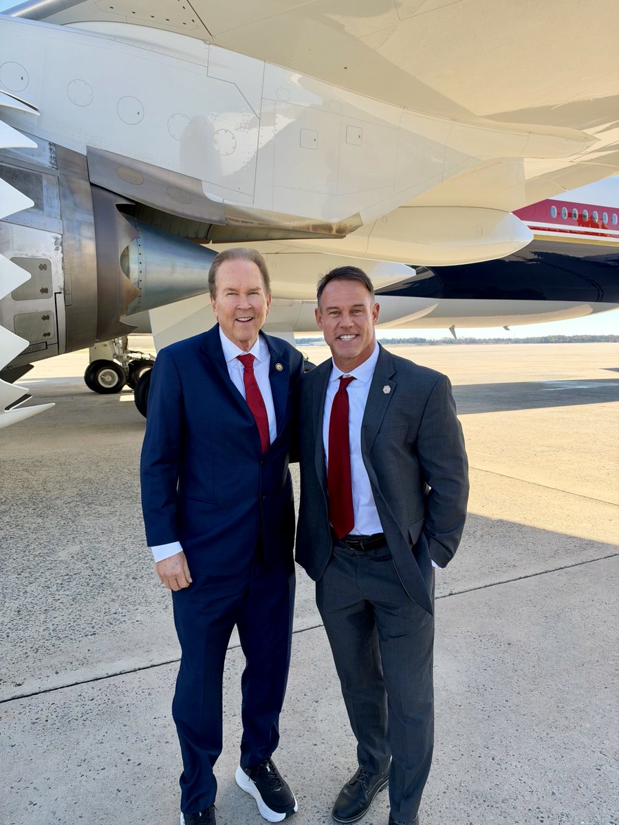Two men in suits pose together on an airport tarmac in front of a large airplane. No text is visible.
