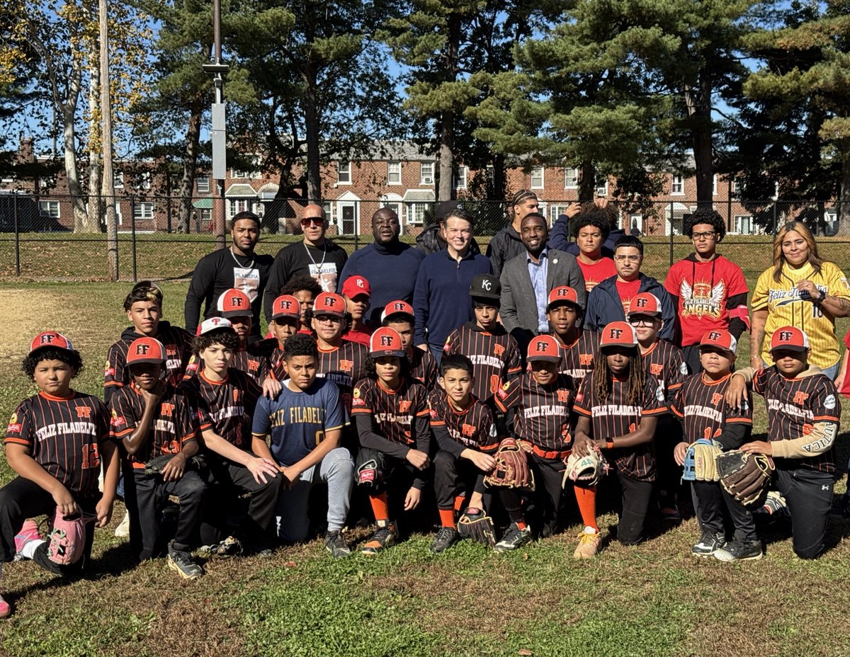 A youth baseball team and several adults pose for a group photo on a sunny field. The players wear matching black-and-orange uniforms and caps, and some hold gloves. There is text on the uniforms and caps.