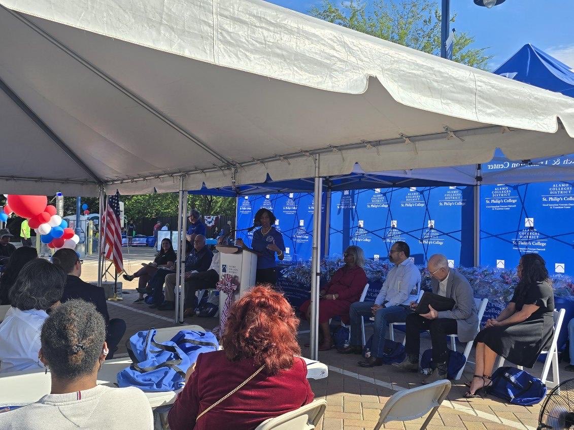An outdoor event under a large white canopy. A woman speaks at a podium while several people sit behind her, and an audience faces the stage. Blue branded backdrops, balloons, and an American flag are visible. There is text on the backdrop and podium.
