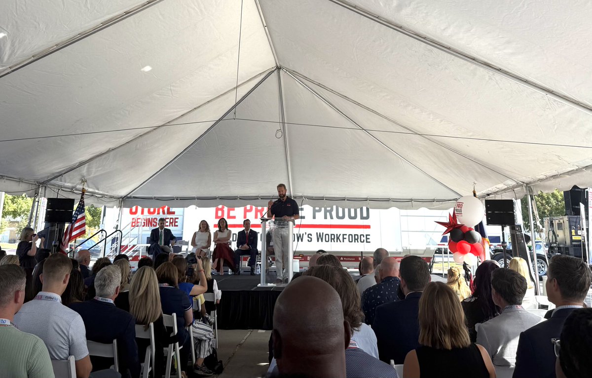 A man speaks at a clear podium on a stage beneath a large white tent, with several people seated behind him and an audience seated in front. A banner, an American flag, speakers, and red, white, and black balloons are also visible. There is text on the banner.