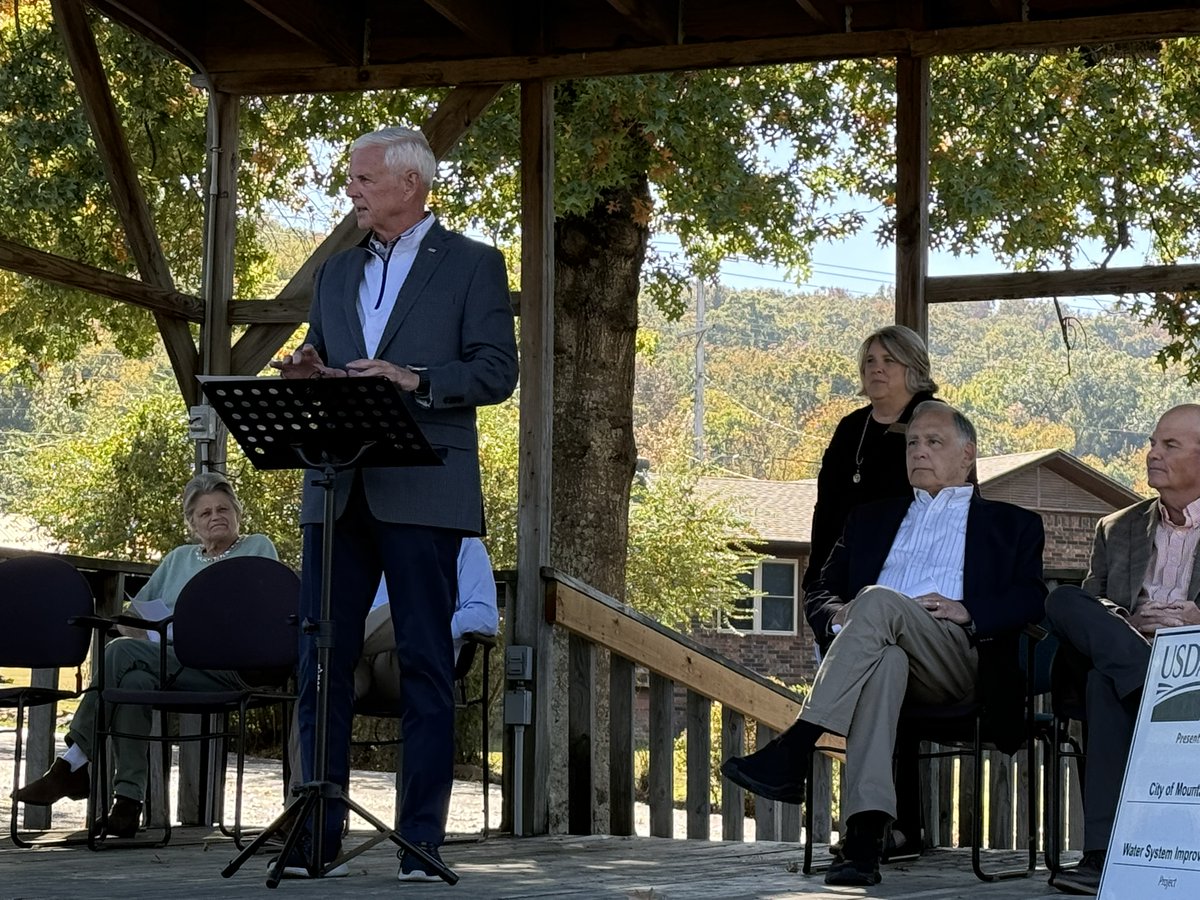An outdoor gathering under a wooden pavilion. An older man in a suit speaks at a lectern while several people sit nearby. Trees and a building are visible behind them, and a sign with text is partly visible at the right edge.