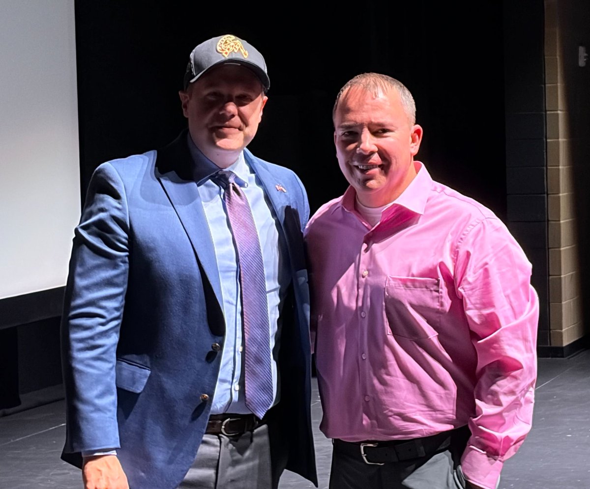 Two men stand side by side indoors, posing for a photograph against a dark background. One wears a suit and baseball cap, and the other wears a pink shirt. There is text on the cap.