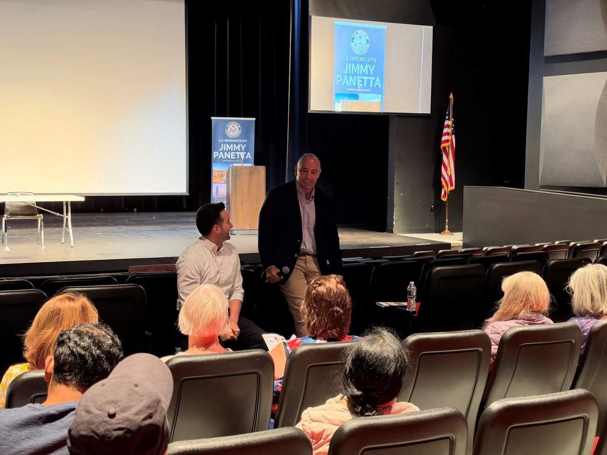 A man holding a microphone stands near seated audience members in an auditorium, beside another seated man. A stage with a podium, projection screens, campaign banners, and an American flag is behind them. There is text on the banners and screens.
