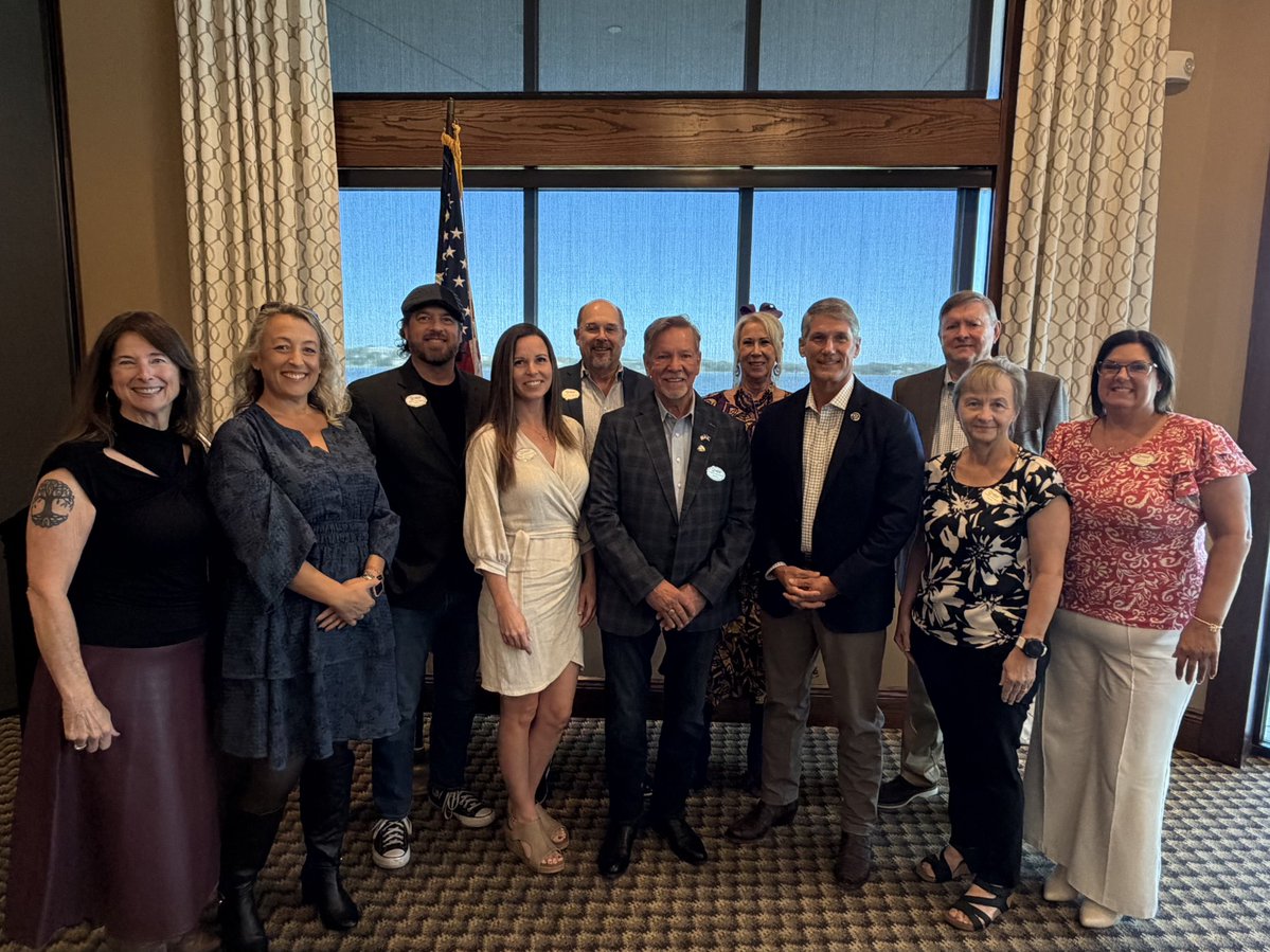 A group of adults pose for a photo in a room with large windows and curtains. An American flag stands behind them. Some people wear small badges with text.