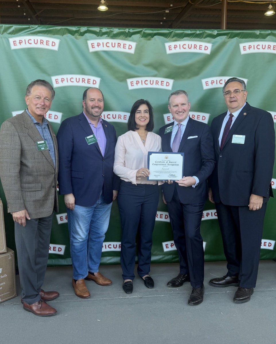 Five adults pose for a group photo in front of a green event backdrop. Two people hold a certificate. There is text on the backdrop, badges, and certificate.