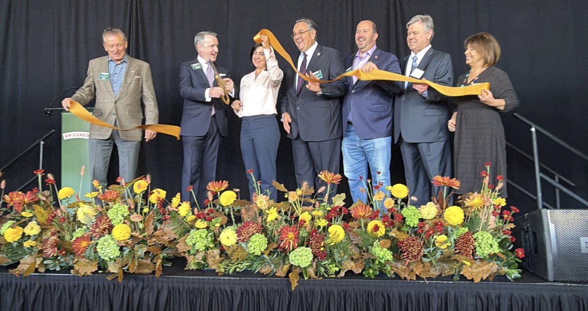 A group of seven people stand on a stage and cut a long orange ribbon during a ribbon-cutting ceremony. A podium and a large arrangement of colorful flowers are also visible. There is text on a sign attached to the podium.