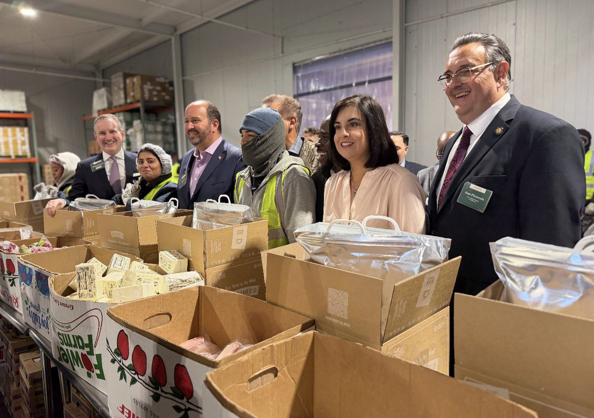A group of people stand together in a warehouse behind open boxes and bags of food supplies, appearing to take part in a food-packing or distribution event. There is visible text on packaging and name badges.