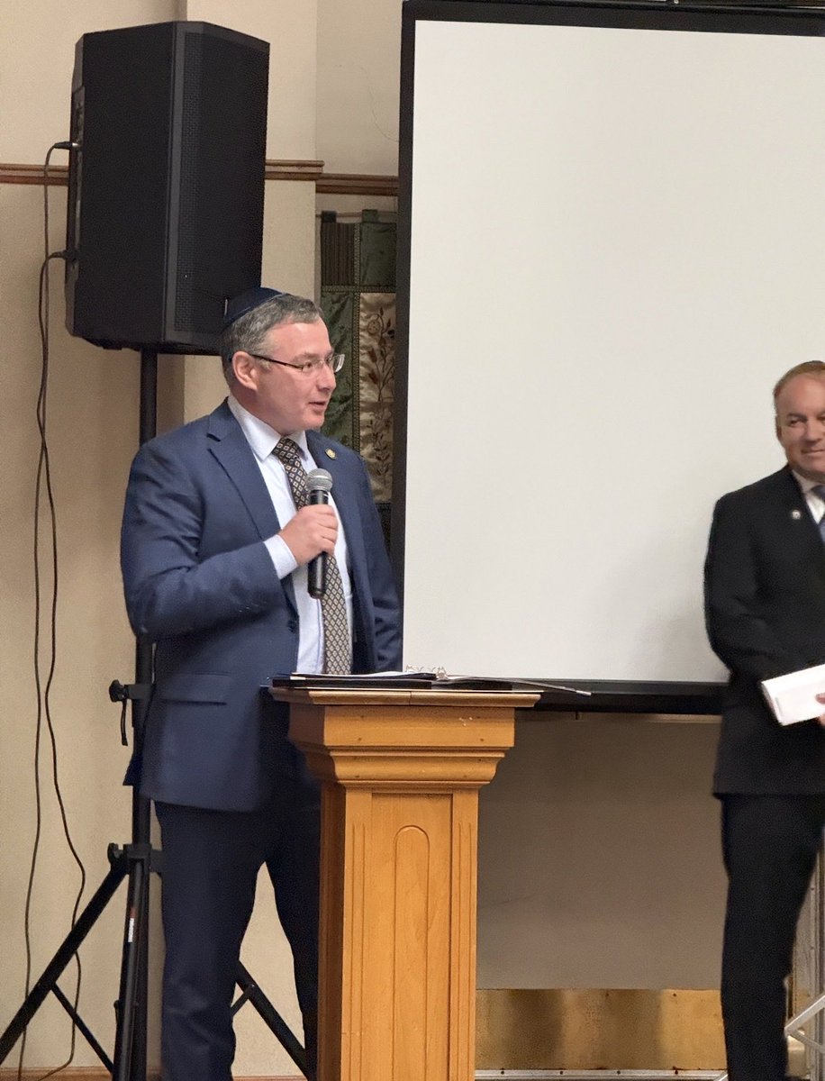 Two men in suits stand at the front of a room. One speaks into a microphone behind a wooden lectern, with a large projection screen and speaker nearby. There is some text visible on papers, but it is not legible.