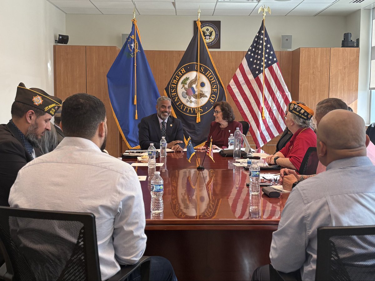 A group of people sit around a conference table during a meeting in an office, with several flags and a government seal displayed behind them. There is text on the flags, seal, and some clothing.