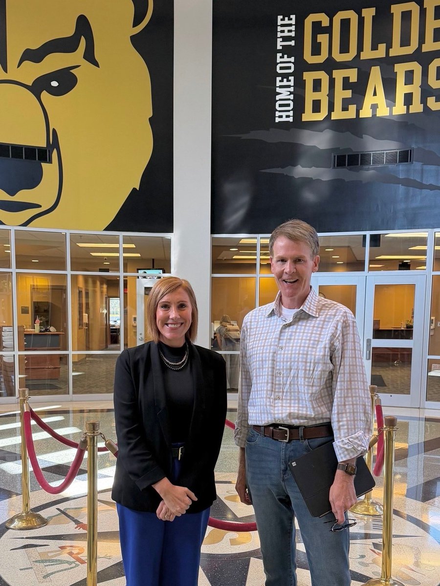 A woman and a man pose for a photo in a school lobby. Behind them are glass-walled rooms, red velvet ropes on gold stanchions, and large black-and-gold school mascot and team signage. Text is visible on the signage.