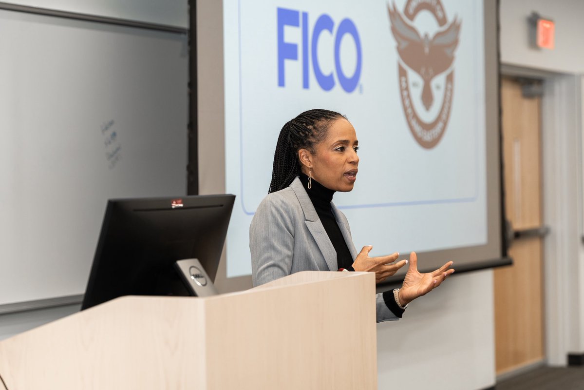 A woman speaks and gestures at a lectern in a classroom or lecture hall. A computer monitor and a projected slide with a logo and text are visible behind her.
