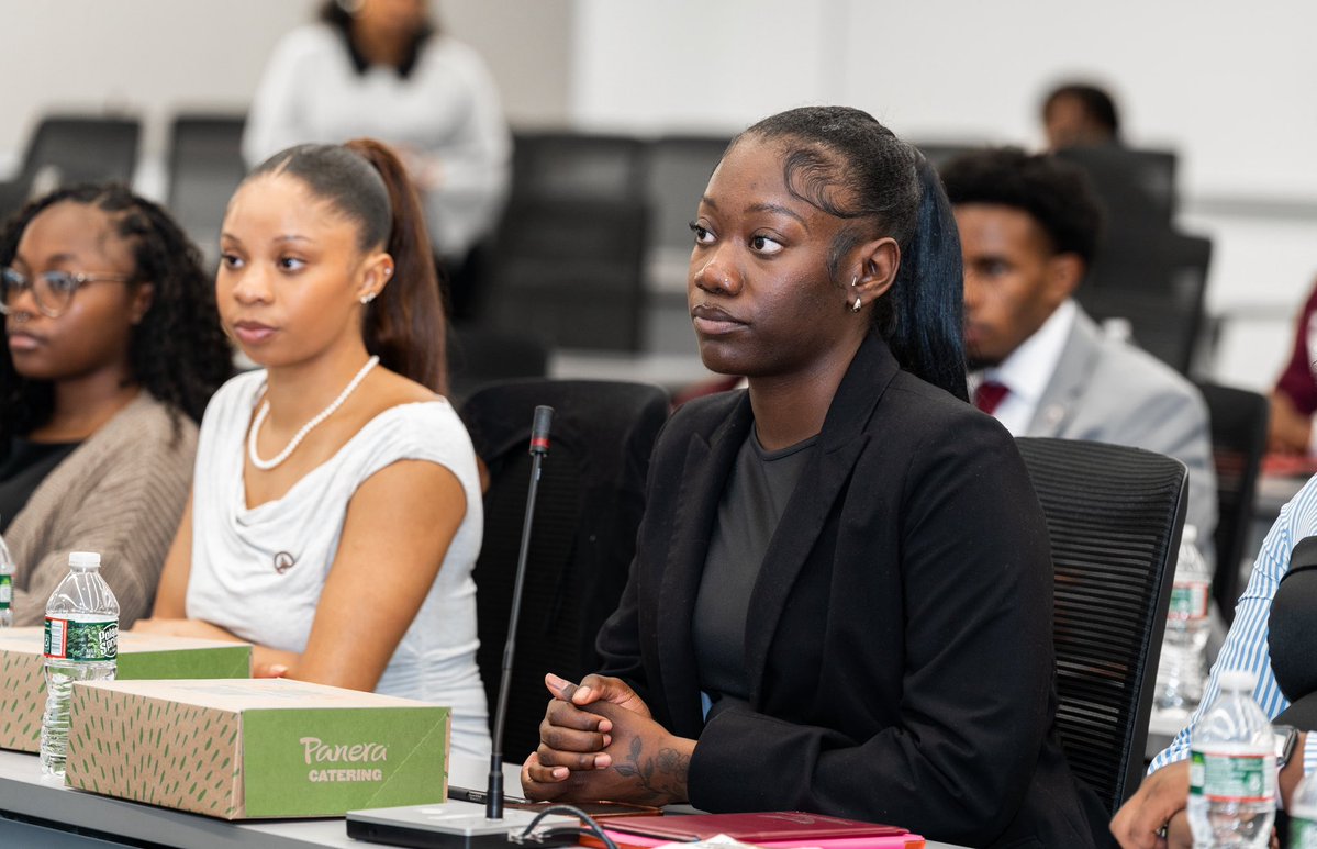 People sit at a conference table in a meeting or classroom, listening attentively. There is visible text on a catering box and drink labels.