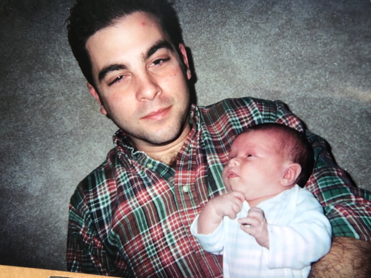A man in a plaid shirt cradles a newborn baby in a white outfit. They are seated against a gray textured background. No text is visible.