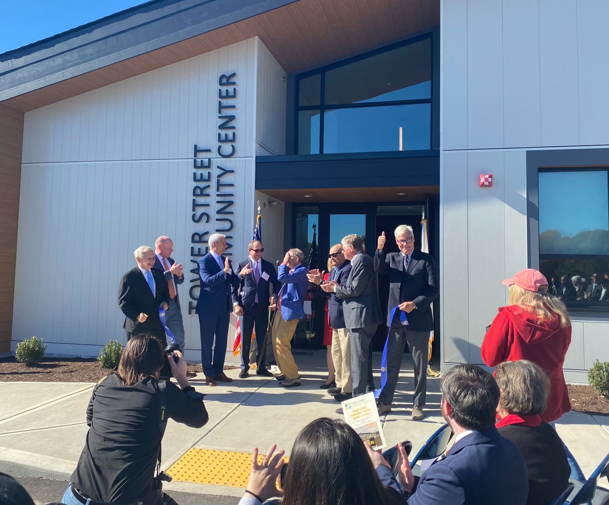 A group of people gathers outside a modern community center for a ribbon-cutting or opening ceremony. Several people near the entrance applaud, while others watch and take photographs. Text is visible on the building and on a small sign.